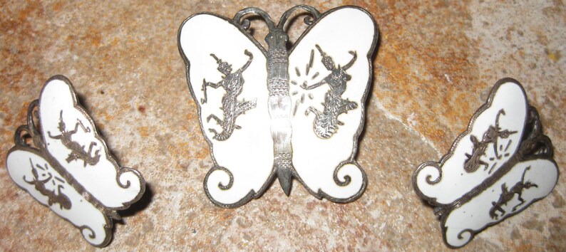 May include: A set of three silver butterfly brooches with white enamel. Each brooch features a stylized butterfly with intricate details and silhouettes of figures.