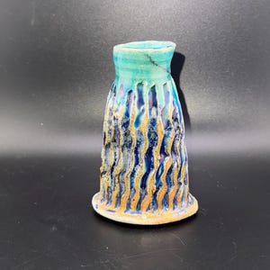 Puede incluir: Un pequeño jarrón de cerámica azul y verde con un diseño texturizado y ondulado. El jarrón tiene una base ancha y un cuello estrecho.