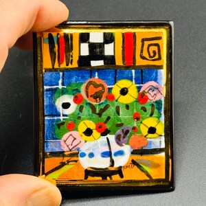 May include: A colorful ceramic tile with a painting of a vase of flowers. The vase is white with blue and white accents. The flowers are yellow, red, and pink. The background is a blue and white checkered pattern. The tile is framed in gold.