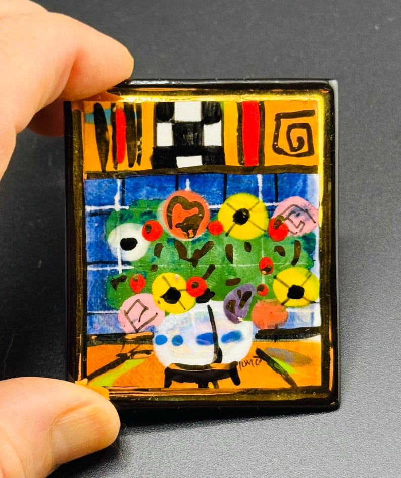 May include: A colorful ceramic tile with a painting of a vase of flowers. The vase is white with blue and white accents. The flowers are yellow, red, and pink. The background is a blue and white checkered pattern. The tile is framed in gold.
