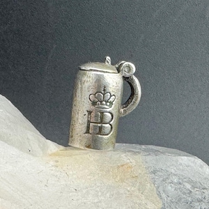 May include: A small silver-toned metal charm shaped like a beer stein with a hinged lid and a handle. The front of the stein features an embossed crown symbol positioned directly above the capital letters HB in a classic serif font style.
