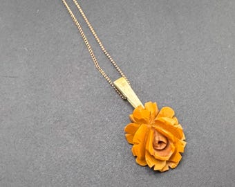 May include: A gold-toned necklace featuring a carved orange rose pendant. The rose is intricately detailed, with visible petals and a central bud. The pendant is attached to a rectangular gold-toned bar, which is connected to a delicate chain.