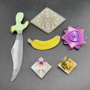 May include: A collection of six decorative items including a light green and silver knife, a diamond-shaped gray brooch with rhinestones, a yellow banana-shaped pin, a purple star-shaped object, and two small diamond-shaped pins with floral designs.