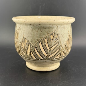 May include: A ceramic planter with a light green speckled glaze. The planter has a repeating leaf design carved into the surface, with a cream-coloured interior visible through the cutouts. The base and rim are a lighter cream colour.