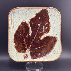 May include: A square ceramic plate with rounded corners featuring a glossy dark brown fig leaf imprint centered on a speckled light cream and pale green background. The edges of the plate are finished in a warm brown tone, highlighting the texture.