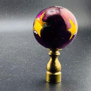 May include: A purple glass lamp finial with gold accents. The finial is round and has a decorative design of yellow and pink shapes.