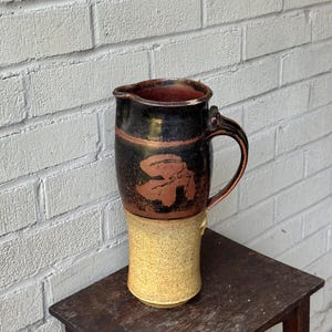 May include: A black and brown ceramic pitcher with a stylized leaf design. The pitcher has a wide mouth and a handle. The bottom of the pitcher is a light yellow color.
