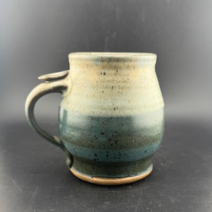 Puede incluir: Una taza de cerámica hecha a mano con un diseño único. La taza presenta un degradado de colores, pasando de un beige claro en la parte superior a un verde azulado oscuro en la base. Tiene un asa resistente y una pequeña pestaña decorativa cerca del borde. La taza mide unos 10 cm de alto.