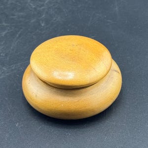 May include: A small, round wooden container with a lid. The container and lid are both a light brown color, with a smooth, polished finish. The lid sits atop the container, creating a layered effect.