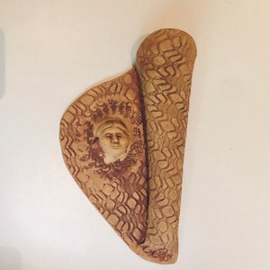 May include: A ceramic wall sculpture with a textured surface and a carved face in the centre. The sculpture is in shades of brown and beige.