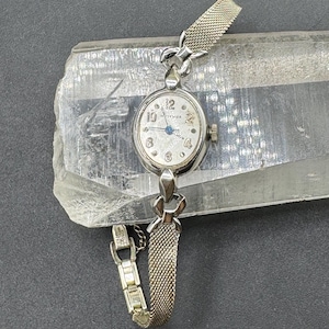 May include: A silver-toned wristwatch with an oval face, featuring a white dial with Arabic numerals, a blue second hand, and a fine mesh metal band. The watch case is connected to the strap by decorative metal links, creating a cohesive design.