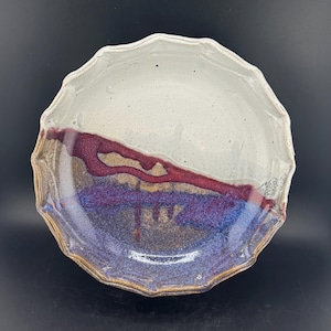 Puede incluir: Un cuenco de cerámica hecho a mano con borde festoneado. El cuenco presenta una mitad superior blanca, una banda roja y morada, y una sección inferior azul y marrón. El borde del cuenco tiene una superficie texturizada e irregular.