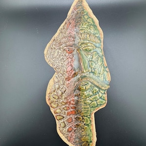 以下が含まれることがあります： 緑と赤の釉薬をかけた顔のセラミック壁彫刻。顔は様式化されており、表面にテクスチャがあります。