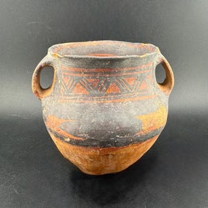 May include: A handmade, ancient-style pottery vessel with two handles. The pot features a dark gray body with orange and black geometric patterns and bands. The vessel has a rounded base and a slightly flared rim, showcasing a rustic aesthetic.