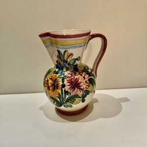 May include: A white ceramic pitcher with a red rim and a floral design in shades of yellow, pink, blue, and green. The pitcher has a handle and a round body.