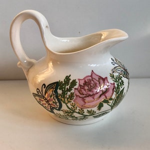 May include: Cream-colored ceramic pitcher with a curved handle and spout. The pitcher is decorated with a pink rose, butterflies, and green foliage with gold accents. The pitcher has a rounded body.