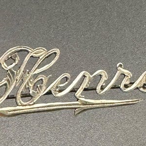 May include: Silver brooch with the name "Bennir" written in a decorative script.