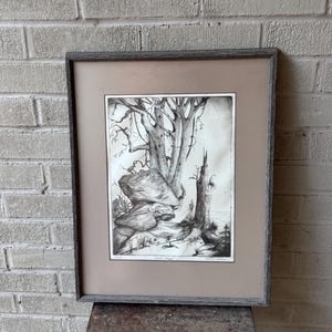 May include: Black and white framed artwork depicting a forest scene with trees, rocks, and a small stream. The artwork is in a gray wooden frame with a tan mat. The artwork is signed in the bottom right corner.