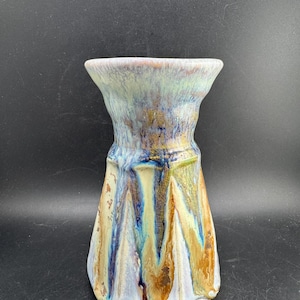 May include: A ceramic vase featuring a flared rim and a tapered body with a reactive glaze finish. The surface displays a blend of cream, light blue, and earthy brown tones, with vertical drip patterns cascading down the textured exterior walls.