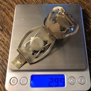 Puede incluir: Un brazalete plateado con bisagras, dise&ntilde;os calados y un elemento central circular. El brazalete est&aacute; sobre una balanza digital, que marca 29,9 gramos. El fondo es una superficie de madera.
