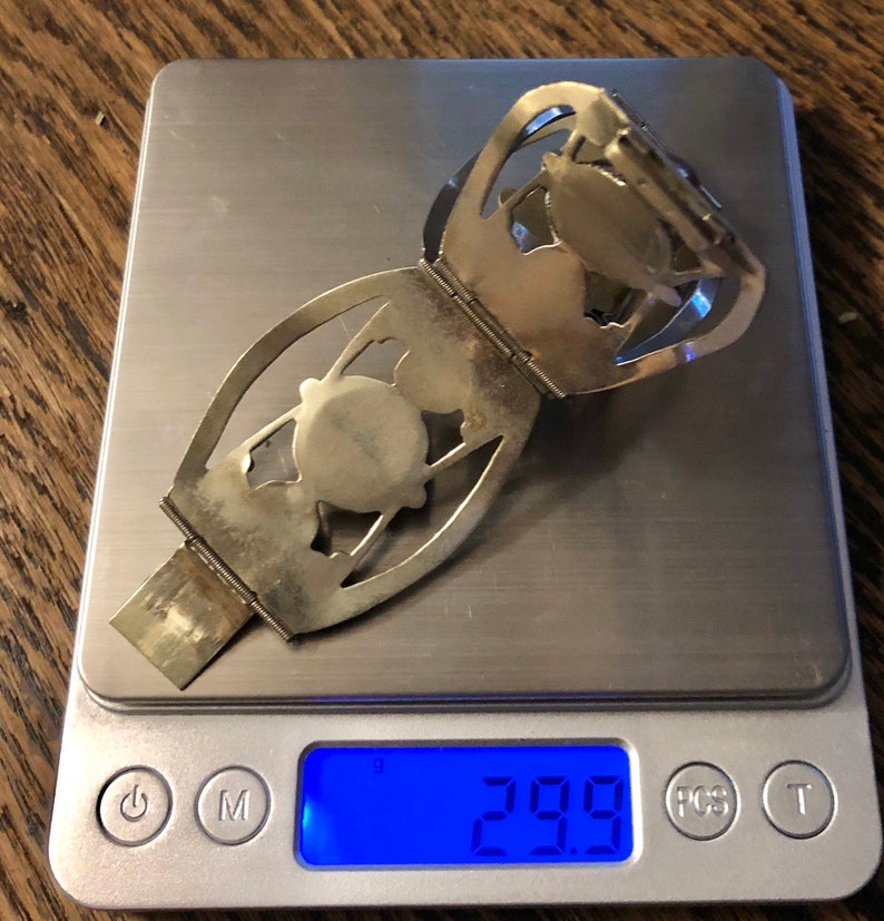Puede incluir: Un brazalete plateado con bisagras, dise&ntilde;os calados y un elemento central circular. El brazalete est&aacute; sobre una balanza digital, que marca 29,9 gramos. El fondo es una superficie de madera.