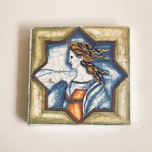 May include: A ceramic tile with a blue and white starburst design. The centre of the tile features a painting of a woman with long, flowing hair, wearing a blue and orange dress. The tile has a distressed, aged look.