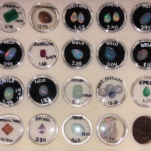 Pode incluir: Uma coleção de pedras preciosas expostas em pequenos recipientes redondos. As pedras preciosas são etiquetadas com seus nomes e pesos, incluindo esmeralda, pedra da lua estrelada, opala welo, rubelita, água, safira estrelada e topázio imperial.