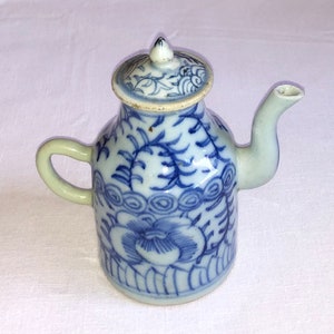 Puede incluir: Una pequeña tetera de porcelana azul y blanca con tapa y diseño floral. La tetera tiene un pico y una asa.
