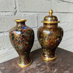 May include: Two cloisonné vases with intricate floral patterns in shades of red, gold, blue, and green. The vases are on a dark wooden surface.