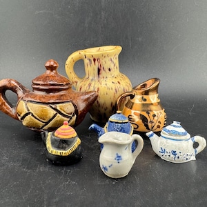 Puede incluir: Una colección de teteras y jarras de cerámica en varios tamaños y diseños. Las teteras presentan patrones marrones, azules y blancos, mientras que las jarras tienen acabados dorados moteados y ornamentados. Los artículos están dispuestos sobre una superficie oscura.