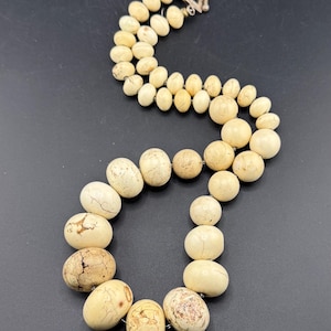 Op de afbeelding: Crèmekleurige kralenketting met een zilveren sluiting. De ketting heeft een dubbele streng van ovale en ronde kralen met een natuurlijke, getextureerde uitstraling. De kralen variëren in grootte en hebben bruine en beige markeringen.