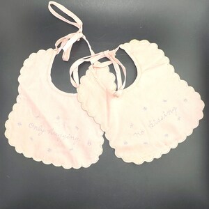 Könnte beinhalten: Zwei blassrosa Baby-Lätzchen mit gewelltem Rand und Stoffbändern. Ein Lätzchen ist mit dem gestickten Text "only hugging" und das andere mit "no kissing" versehen, beide akzentuiert durch kleine, verstreute violette Blumenstickereien.