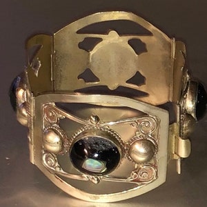 Puede incluir: Una pulsera de metal dorado con intrincados dise&ntilde;os calados y elementos decorativos. La pulsera presenta un panel central rectangular con una piedra grande, oscura y ovalada y piedras redondas m&aacute;s peque&ntilde;as. Tiene un cierre de bisagra.