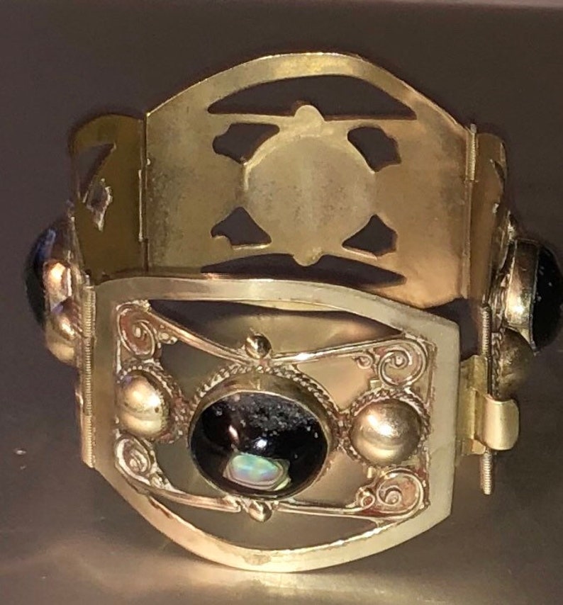 Puede incluir: Una pulsera de metal dorado con intrincados dise&ntilde;os calados y elementos decorativos. La pulsera presenta un panel central rectangular con una piedra grande, oscura y ovalada y piedras redondas m&aacute;s peque&ntilde;as. Tiene un cierre de bisagra.