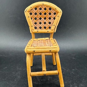 May include: A miniature wooden chair with a woven back and seat. The chair is made of light brown wood and has a curved back. Small metal tacks secure the woven elements to the frame. The chair is a decorative item.