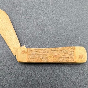 May include: A wooden pocket knife with a brown wood handle and a single blade. The handle has a textured wood grain pattern and a small circular design on the end.