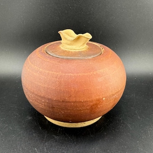 May include: A round, terracotta-colored ceramic jar with a lid. The lid has a unique, ruffled design and is a lighter shade of brown.