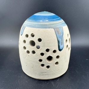 Puede incluir: Un portavelas de cerámica blanca con un esmalte azul y gris. El portavelas tiene forma de cúpula con una serie de pequeños agujeros alrededor de los lados.