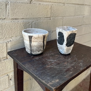 May include: Two ceramic cups with black and white glaze. The cups are sitting on a dark wooden table.