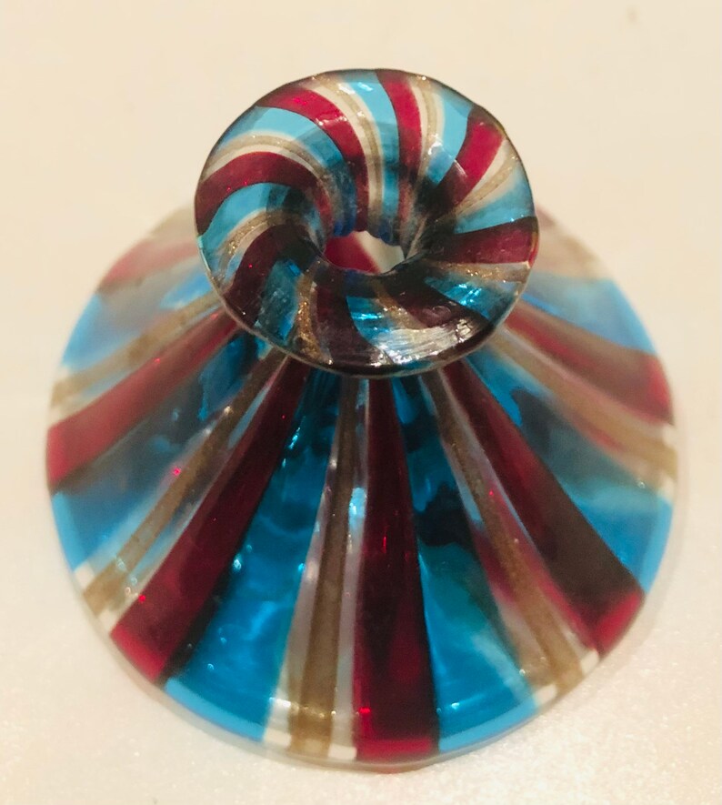 May include: A decorative glass object with a flared base and a rounded top. The design features alternating stripes of blue, red, and gold. The object appears to be a small vase or a unique drinking glass.