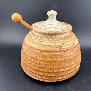 May include: A ceramic honey pot with a lid and a wooden honey dipper. The pot is a warm, earthy color with horizontal bands of brown and orange. The lid is off-white with a textured surface. The wooden dipper rests on the side.
