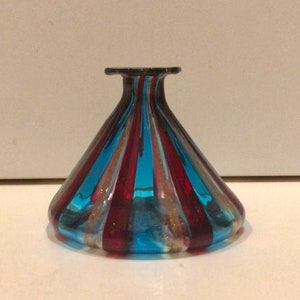 May include: A decorative glass vase with a flared base and a narrow neck. The vase has alternating vertical stripes of blue, red, and gold. The glass is transparent, allowing light to pass through. The vase is likely used for display.