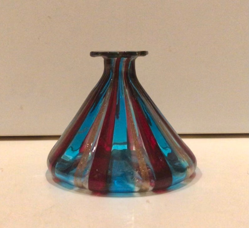 May include: A decorative glass vase with a flared base and a narrow neck. The vase has alternating vertical stripes of blue, red, and gold. The glass is transparent, allowing light to pass through. The vase is likely used for display.