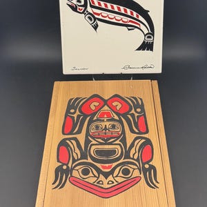 Könnte beinhalten: Eine Holzkiste und eine weiße Fliese mit nordwestamerikanischer Kunst. Die Kiste hat ein rotes, schwarzes und weißes Design. Die Fliese zeigt einen schwarzen Lachs mit roten Akzenten. Das Wort "Salmon" steht auf der Fliese.