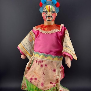 May include: A decorative doll featuring a painted face with red, black, and green patterns. The doll wears a pink tunic, a beige apron with floral motifs, and a blue headpiece adorned with red and blue pom-poms. Green fringe hangs from the waist.