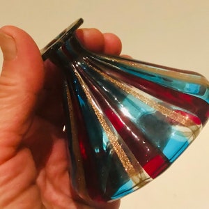 May include: A decorative glass vase with a flared, rounded form. The vase has alternating stripes of red, teal, and blue, with gold glitter accents. The vase is held in a hand, highlighting its unique design and craftsmanship.