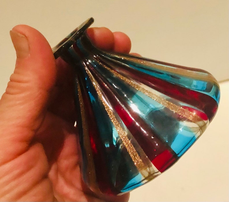 May include: A decorative glass vase with a flared, rounded form. The vase has alternating stripes of red, teal, and blue, with gold glitter accents. The vase is held in a hand, highlighting its unique design and craftsmanship.