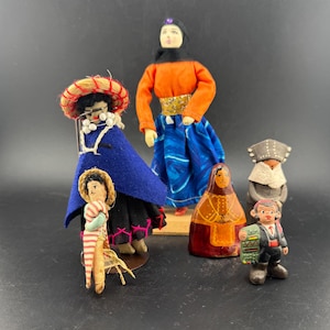 Op de afbeelding: Een verzameling van zes vintage volkskunstpoppen. De poppen zijn gekleed in kleurrijke traditionele kleding, waaronder hoeden, jurken en jassen. De poppen zijn gemaakt van verschillende materialen, waaronder hout, stof en stro. De poppen zijn gerangschikt op een zwart oppervlak.