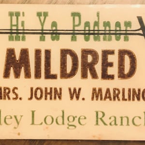 May include: A vintage name tag with the name "Mildred" in brown, block letters. The tag also includes the text "Mrs. John W. Marling" and "Valley Lodge Rancher" in green lettering. The background is a cream color.