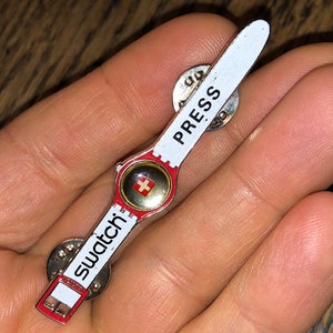 Puede incluir: Un pin de esmalte blanco y rojo con forma de reloj Swatch con la palabra "Swatch" y una bandera suiza en la esfera. La parte posterior del pin dice "Press".
